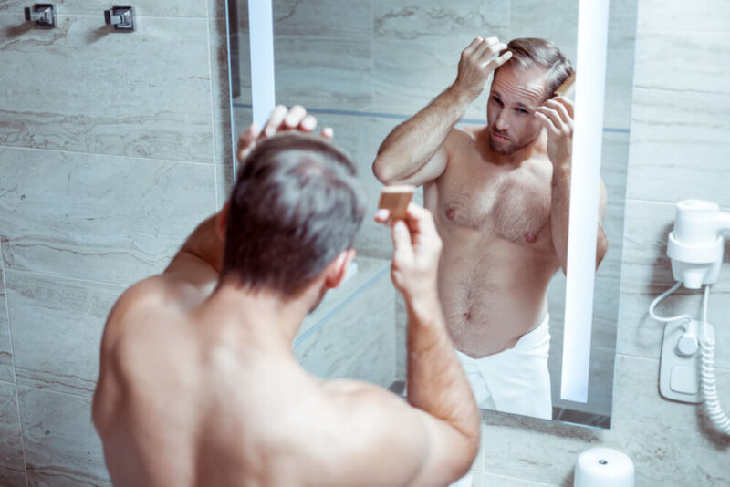 Shitless young white caucasian male in front of the toilet mirror, checking his Norwood 1 hair loss stage